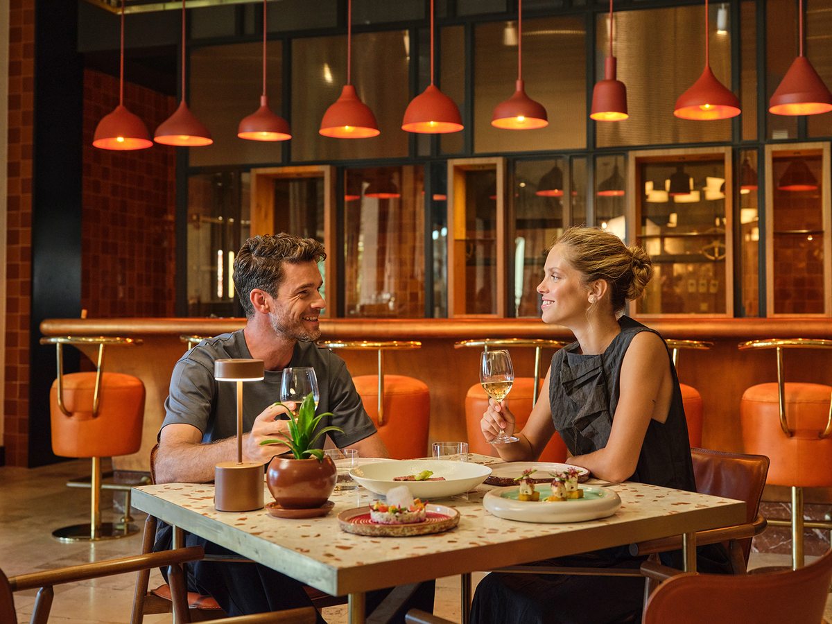 A couple eating at a terrazzo table in a modern restaurant, with a curved bar, orange leather stools and a row of red pendant lights behind them.
