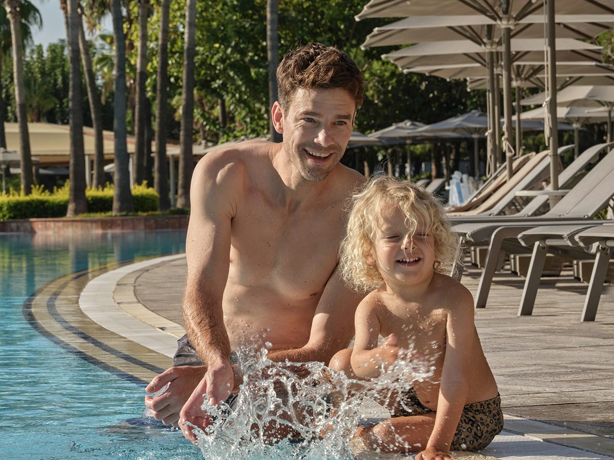 A man and a small child kneeling at the shallow curved edge of a large pool, splashing water, with sun loungers and parasols along the poolside behind.