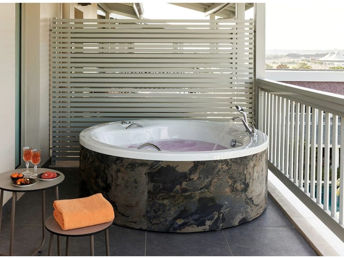 A round stone jacuzzi on a terrace behind a slatted timber screen, with a folded orange towel and a small table set with two drinks and a bowl of fruit.