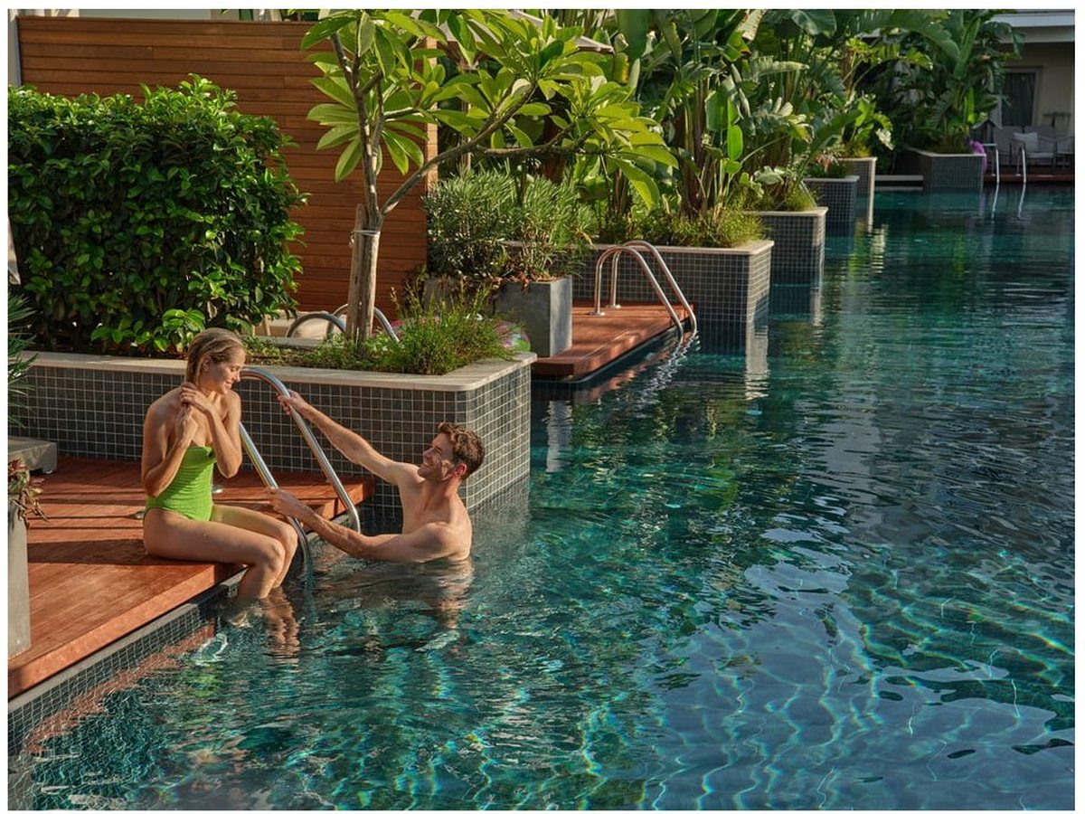 Two people in a long turquoise pool beside a wooden deck, with planters of shrubs and palms and a low tiled wall along the water's edge.