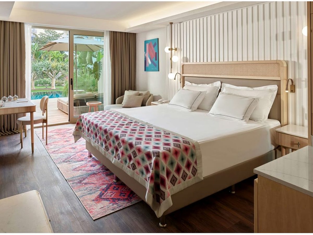 A room with a double bed dressed in white under a red patterned throw, a red kilim rug and sliding glass doors open to a terrace with the pool and gardens beyond.