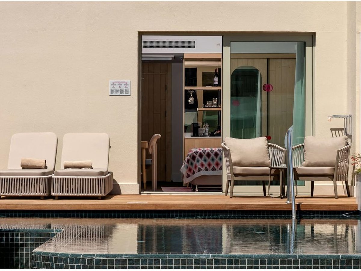 A ground-floor room terrace with two sun loungers and armchairs on wooden decking, its sliding doors open, and a dark-tiled pool running right up to the deck.