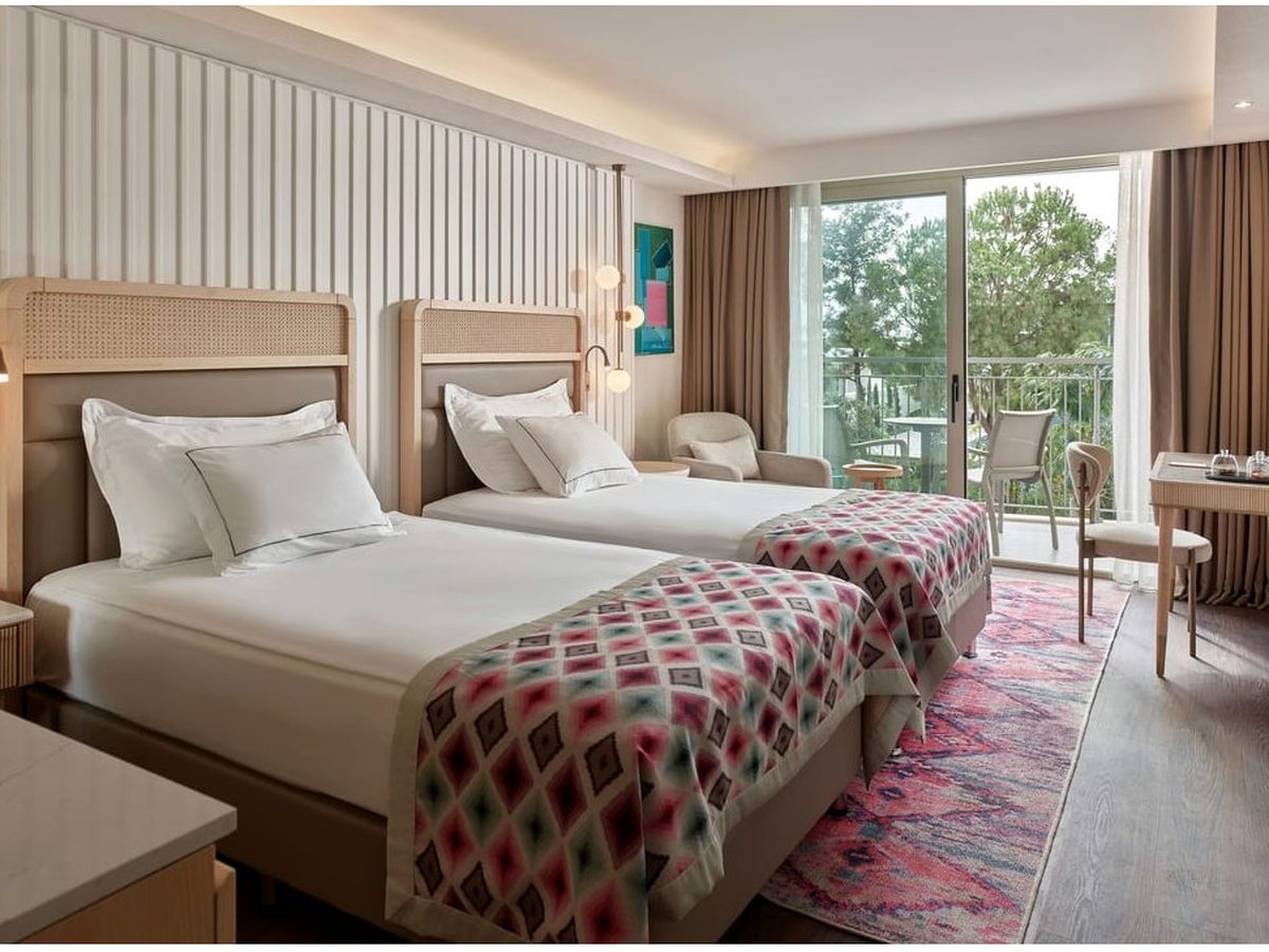 A room with two single beds, cane-panelled headboards, a red patterned throw across each bed and glass doors to a balcony with a table and chairs.