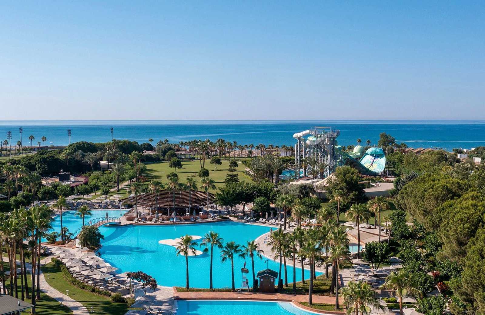 Lara Barut Collection from the air: a large free-form pool ringed by palms and sun loungers, lawns and the aquapark slides behind, and the Mediterranean beyond.