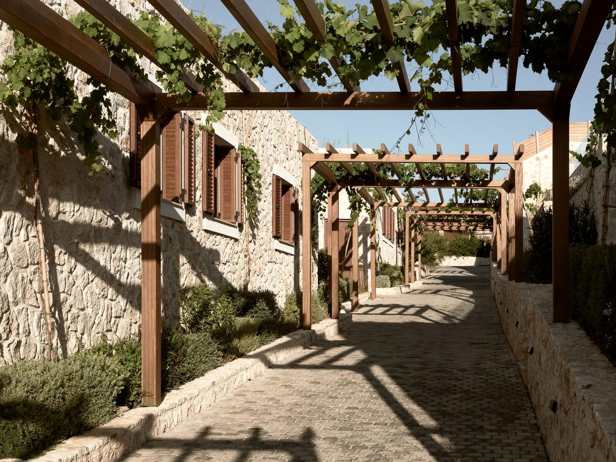 Stone-paved walkway running between rough stone buildings with wooden shutters, under a timber pergola covered in vines.