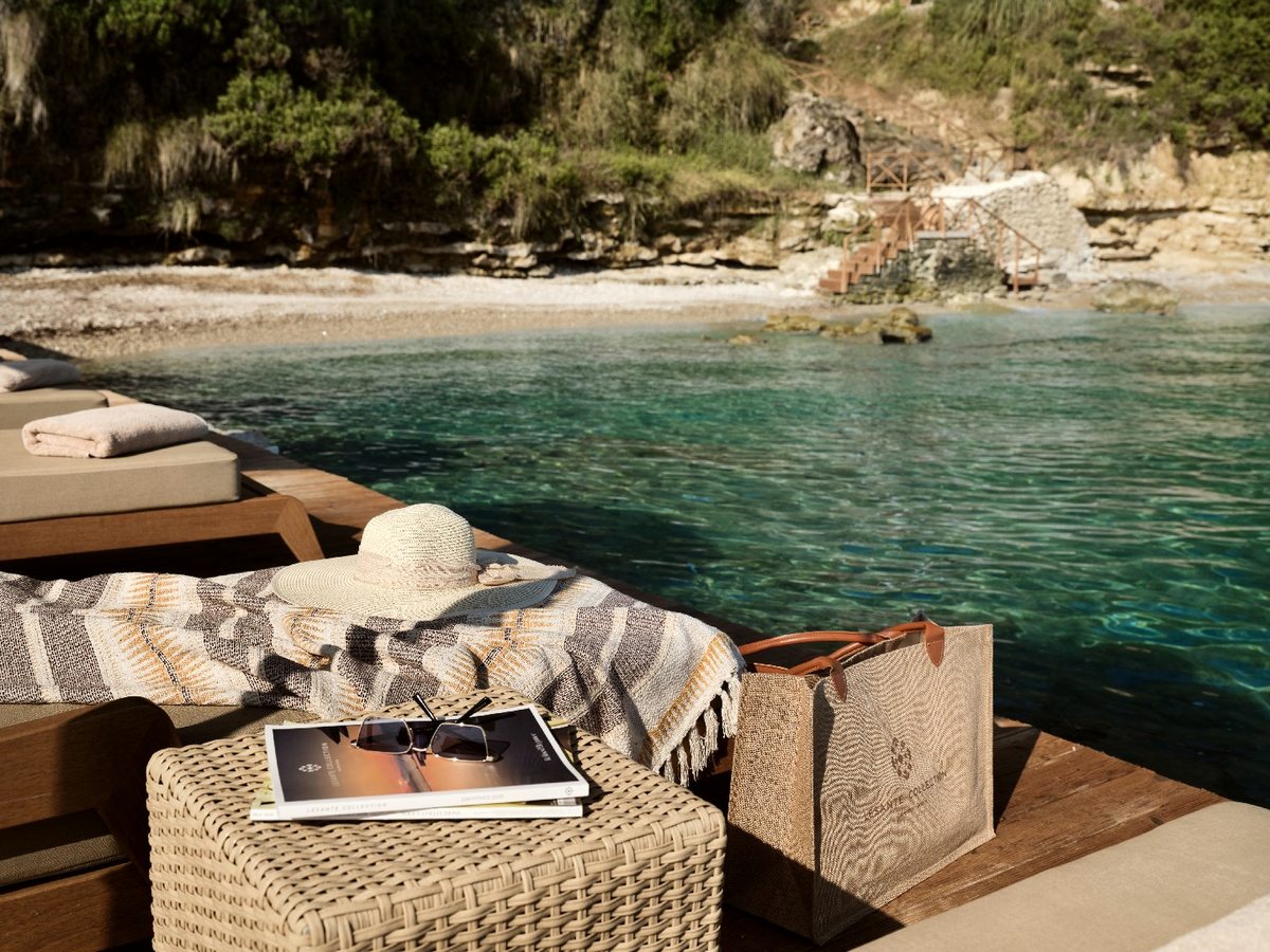 Small pebbled cove with clear shallow water, backed by scrubby cliffs, with a cushioned daybed and a straw hat in the foreground.