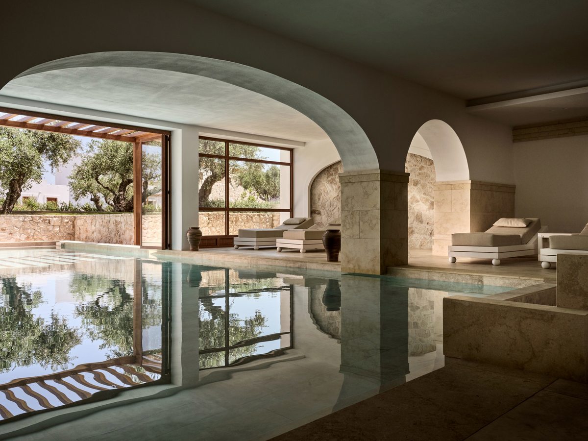 Indoor pool in a stone-arched room with pale plaster walls, cushioned loungers along one side and windows onto an olive grove.