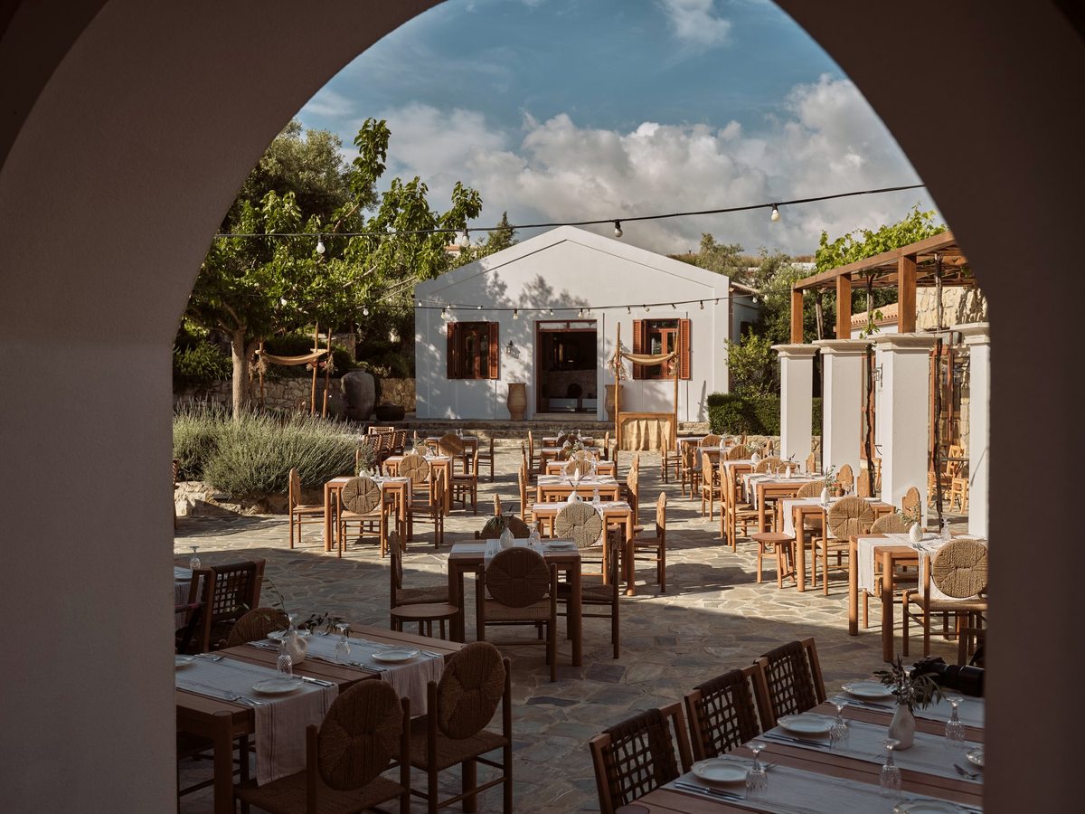 Outdoor courtyard of wooden tables and rush-seated chairs under festoon lights, in front of a white village building with a bell.