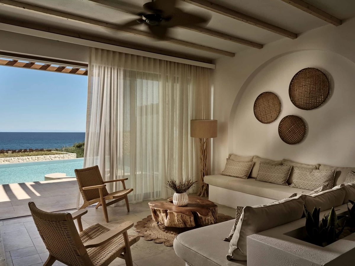 Villa living room with pale sofas, woven wall discs and timber armchairs, opening through full-height doors onto a private pool terrace and the sea.