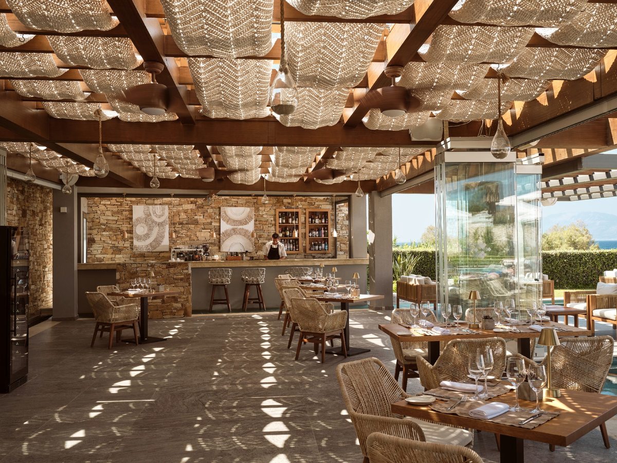 Open-sided restaurant pavilion under a patterned woven pergola, with wicker chairs, wooden tables and a bar counter, opening to the pool and sea.