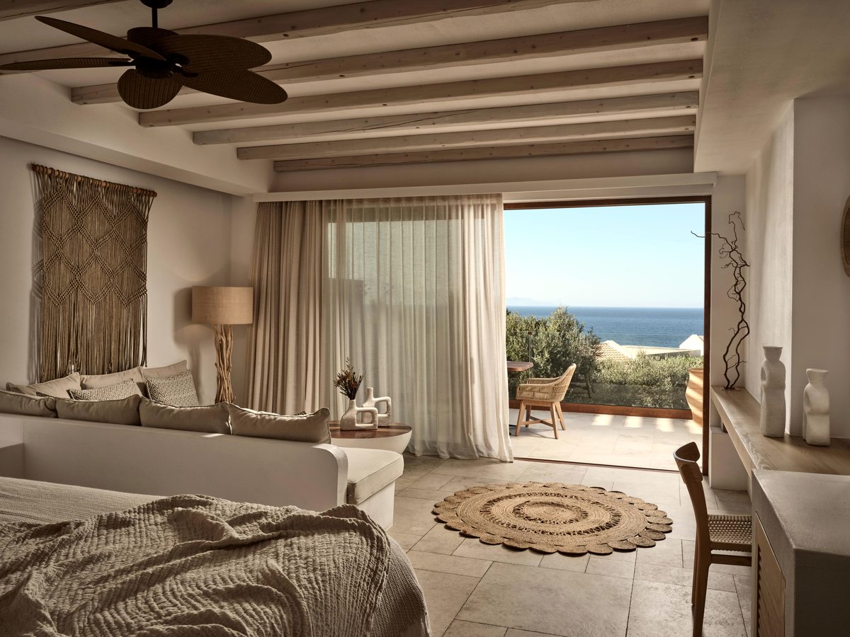 Suite bedroom in cream and sand tones with a beamed ceiling, woven rug and open glass doors onto a terrace with the sea beyond.