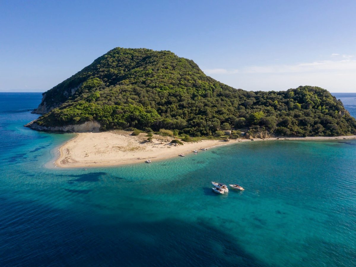Aerial view of a small wooded island joined to a sandy spit, ringed by clear turquoise shallows with a boat moored offshore.