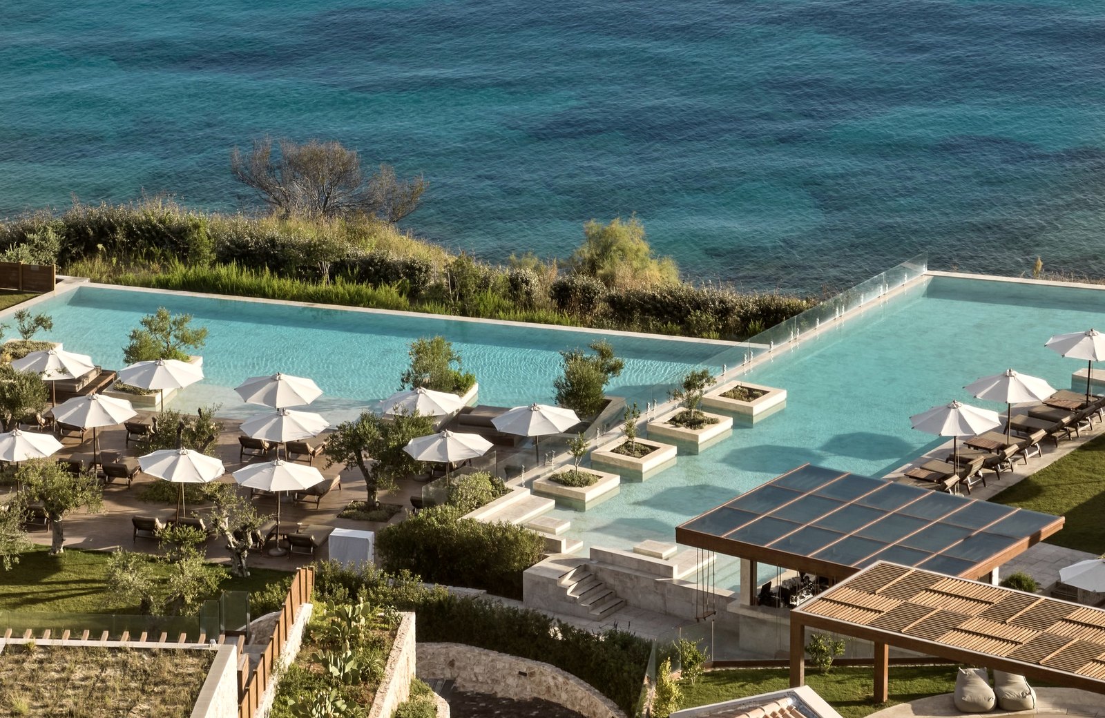 Two of Lesante Cape's turquoise infinity pools on stepped stone terraces above the Ionian Sea, edged with olive trees, sun loungers and cream parasols.