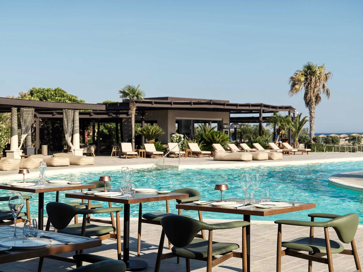 Long dining tables with green and dark chairs set out on a terrace at the edge of a large blue pool, with sunbeds, a pergola and palm trees along the far side.