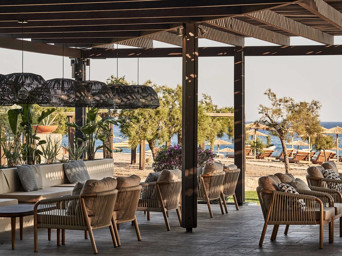 Deep wicker armchairs, sofas and low tables under a timber pergola hung with woven basket lights, opening onto sunbeds, parasols and the sea.