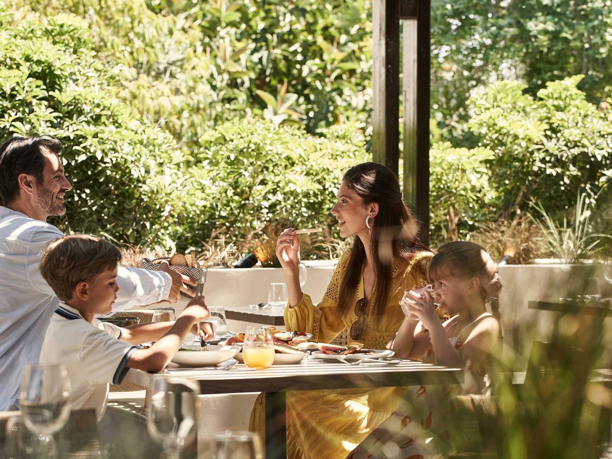 Two parents and two young children sitting at a table of food and orange juice on a shaded open-sided terrace surrounded by green foliage.