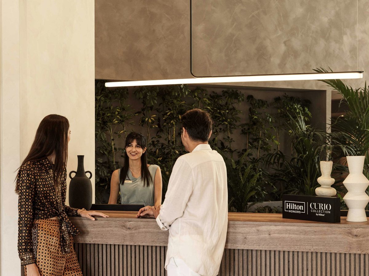 Two guests standing at a slatted timber reception desk talking to a smiling receptionist, with a wall of green planting behind and a Hilton Honors Curio Collection sign on the counter.