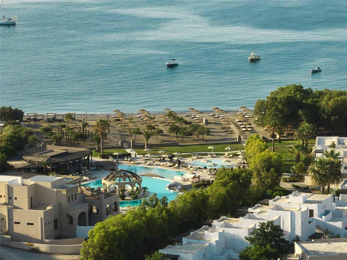 Elevated view over white low-rise resort buildings and a freeform turquoise pool to a long tree-dotted beach lined with straw parasols, and a calm blue bay with small boats at anchor.