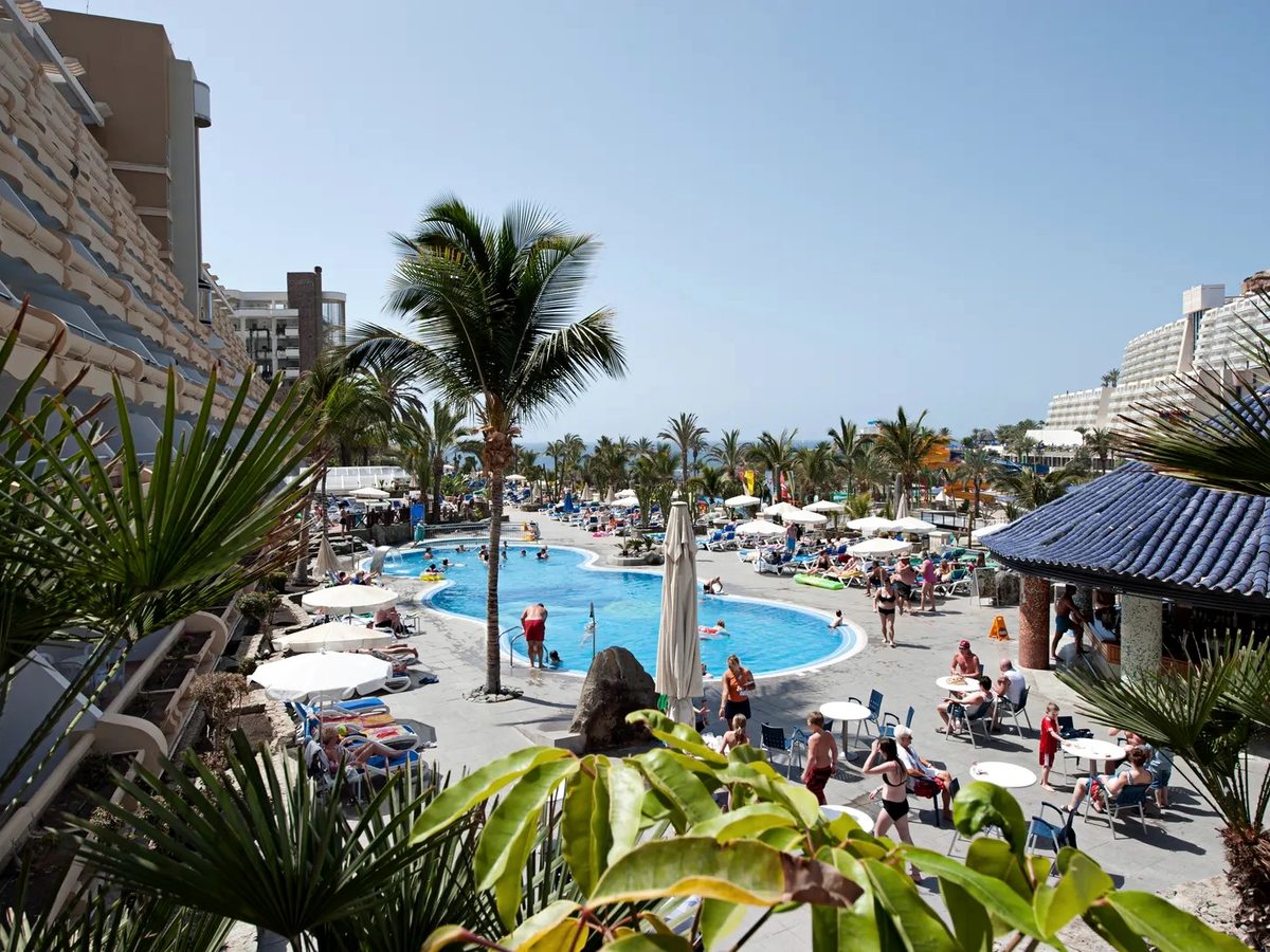 Curved blue pool with families swimming, sun loungers and white parasols around the tiled deck, tall palms, a blue-tiled poolside pavilion and the hotel block along one side.