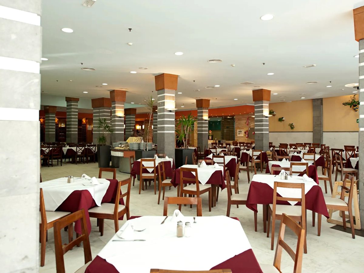 Buffet restaurant dining room with rows of wooden tables laid with white cloths and burgundy napkins, wooden chairs, striped columns and the buffet counters along the far side.