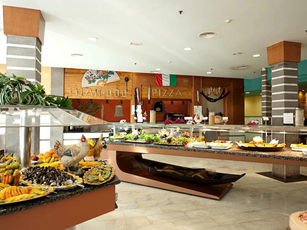 Buffet island of fruit and cold dishes under glass guards, with a pizza counter and its sign on the wall behind.
