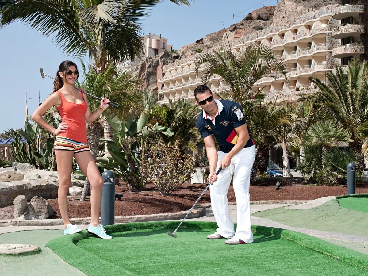 Mini-golf course of artificial-turf greens edged with volcanic rock and palms, two players with putters, and the terraced balconies of the hotel rising behind.
