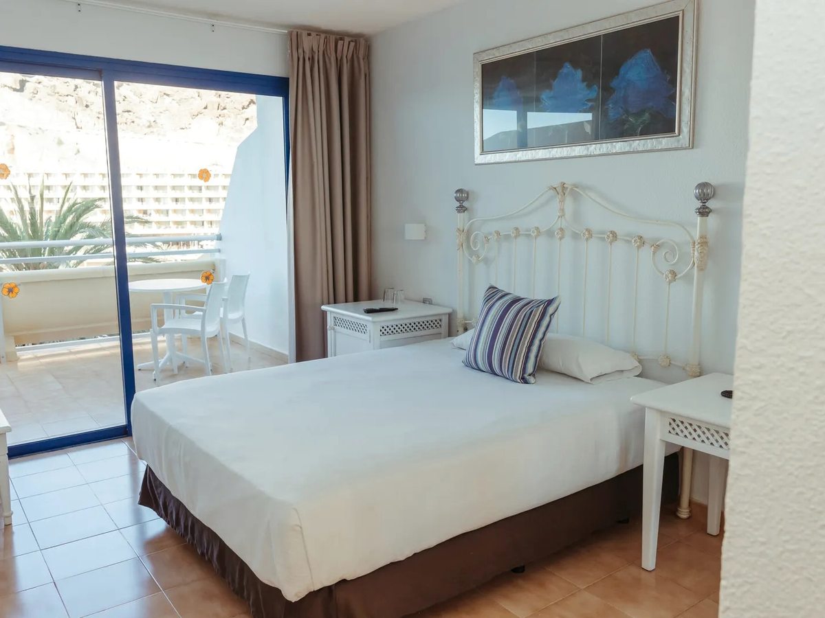 Double room with a white metal bedstead, bedside tables and lamps, framed artwork and sliding glass doors onto a furnished balcony.