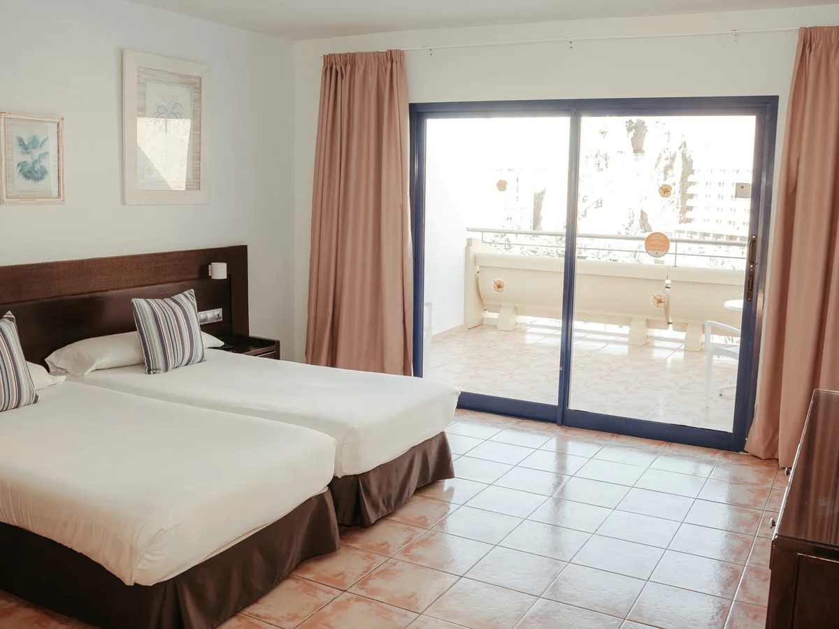 Twin room with two single beds under a shared dark-wood headboard, striped cushions, framed prints and sliding glass doors onto a balcony with a table and chairs.
