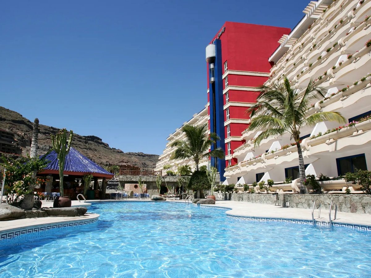 Large curved pool with mosaic-tiled edging and steps, a blue-tiled poolside pavilion to one side, palms, the hotel's white balconied wings and red tower behind and bare mountains beyond.