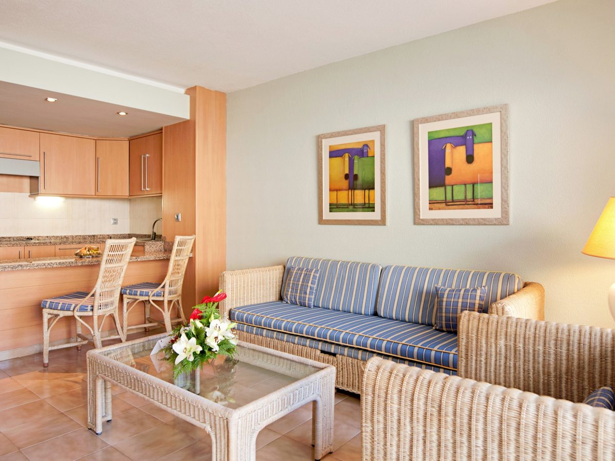 Apartment living room with a blue striped sofa and wicker armchairs around a glass-topped wicker table, framed prints on the wall and an open kitchenette with a breakfast bar and two stools behind.