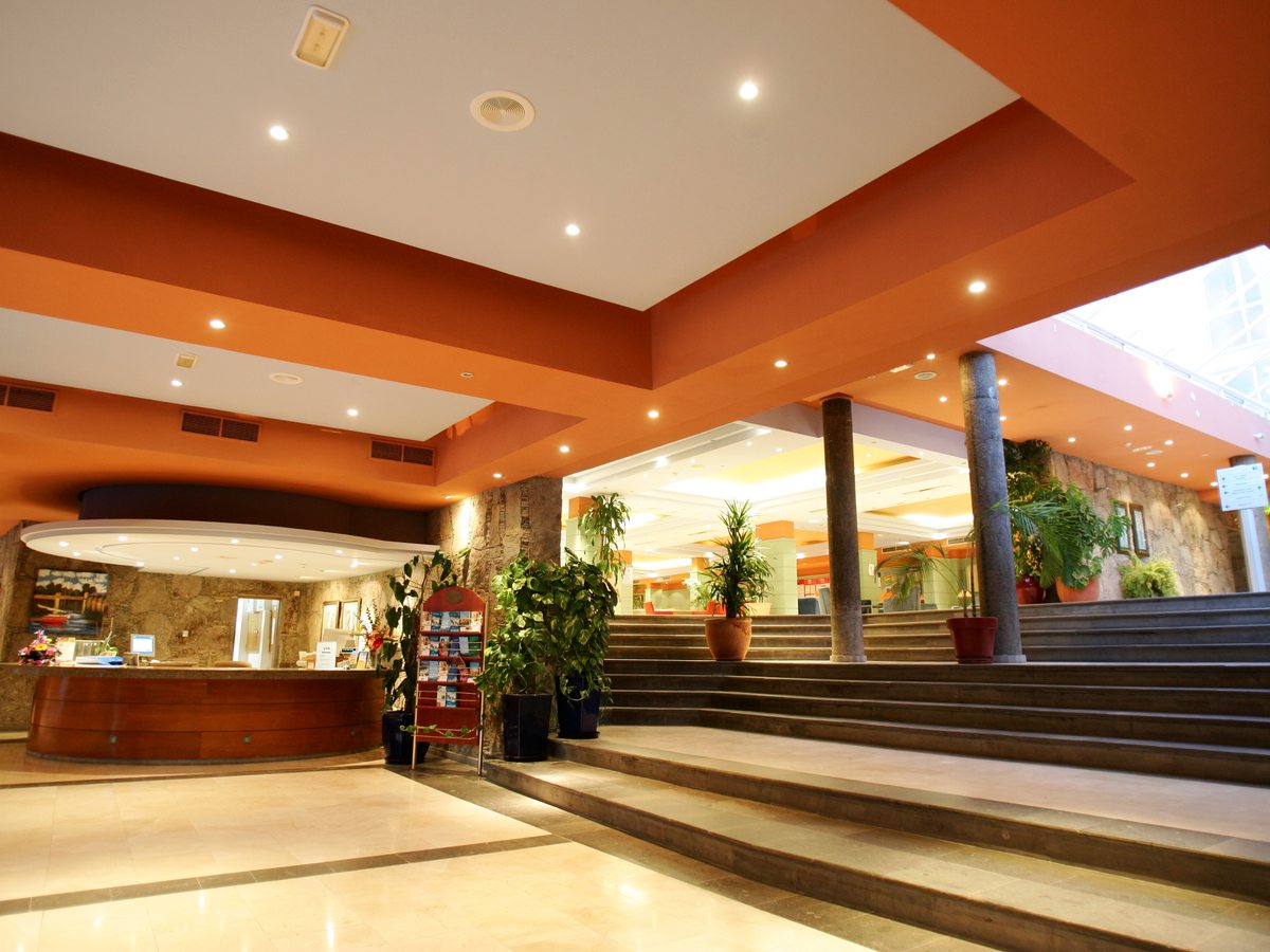 Hotel lobby with a curved reception desk, an orange coffered ceiling, square columns, potted plants and a wide staircase rising to a skylit atrium.