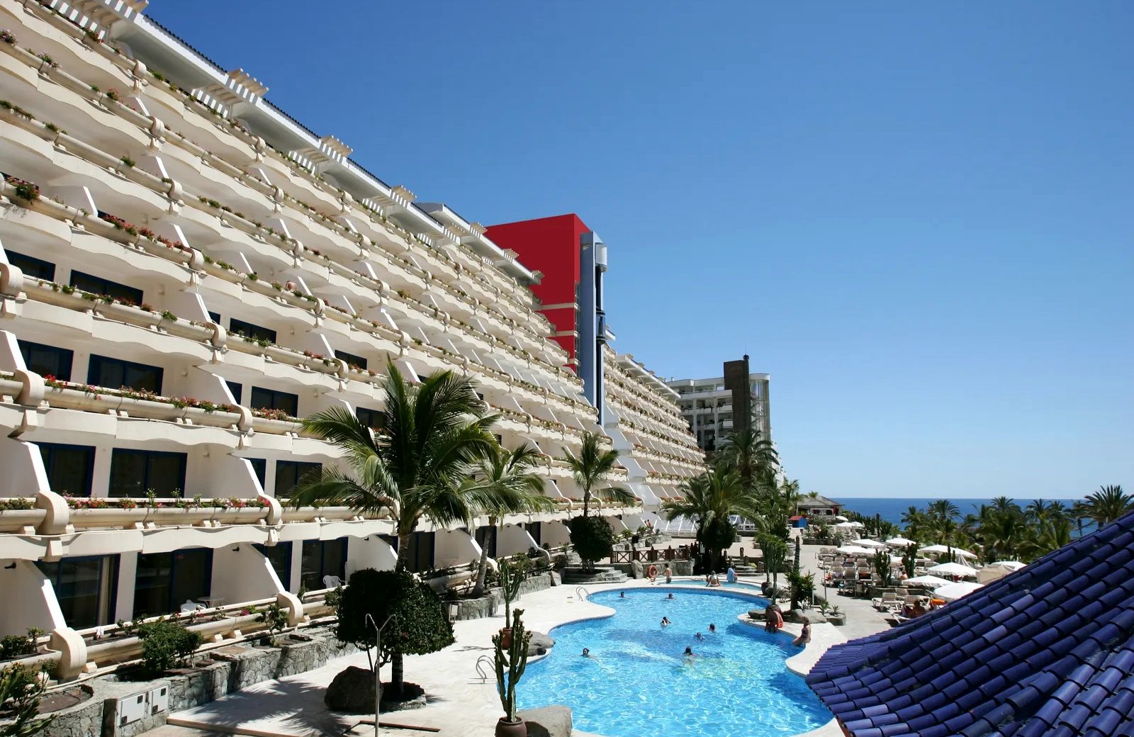 The terraced, flower-lined balconies of Hotel LIVVO Lago Taurito rising above its curved outdoor pool, with palms, sun loungers and the sea beyond.