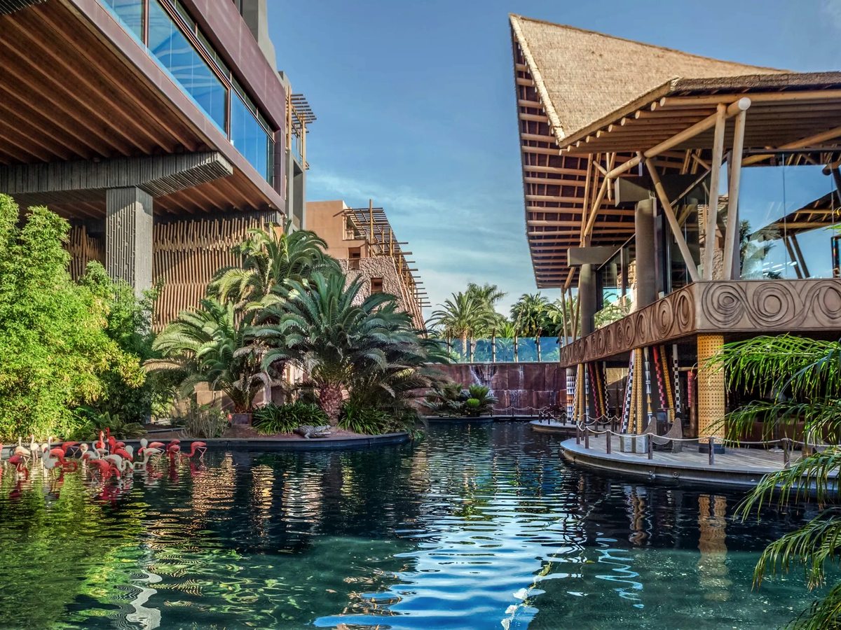 A still green lagoon running between a thatched timber pavilion on one side and a tiered resort building on the other, with palms, bamboo and pink flamingo figures along the water's edge.