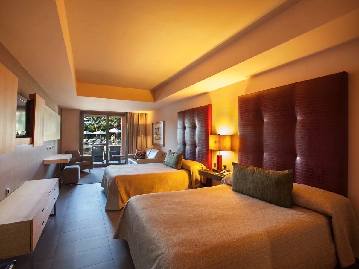 A family room with two queen beds side by side under tall padded headboards, a sofa and armchair seating area, and full-height doors onto a balcony above the pool deck.