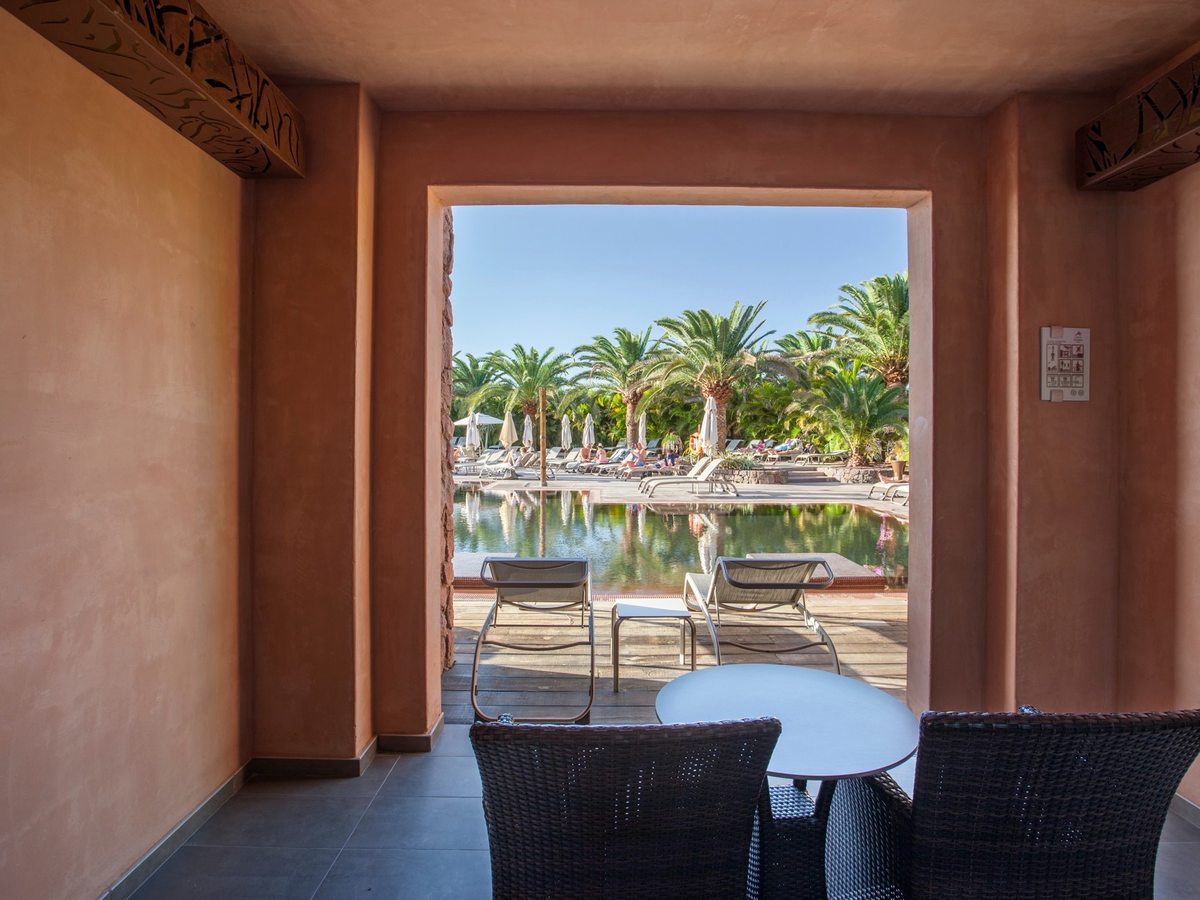 A shaded room terrace with wicker chairs and a round table opening straight onto a timber pool deck, with sun loungers, a still pool and palms beyond.