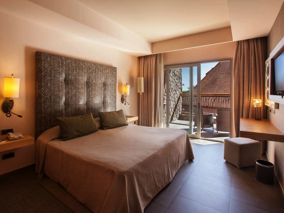 A guest room with a king-size bed against a tall padded headboard, bedside lamps, a desk and a wall-mounted television, with balcony doors open to the gardens.