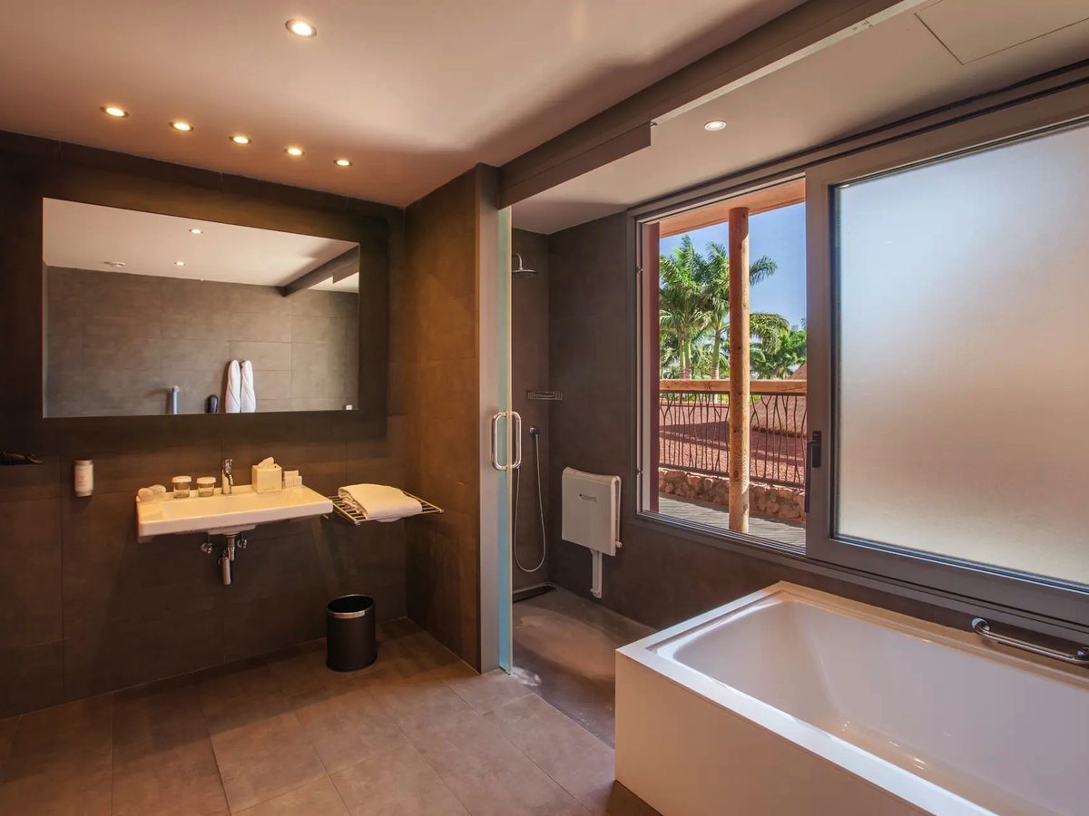 A bathroom with a wide vanity and mirror, a bath under a frosted window and a separate glass-screened shower on dark tiles.