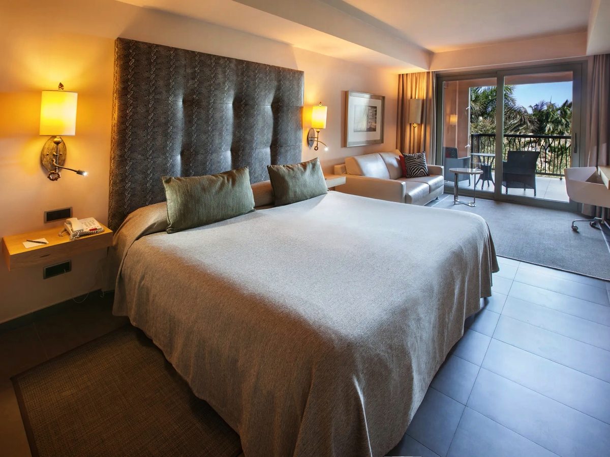 A guest room with a large bed and, beyond it, a sofa and armchair seating area beside sliding doors onto a furnished balcony.