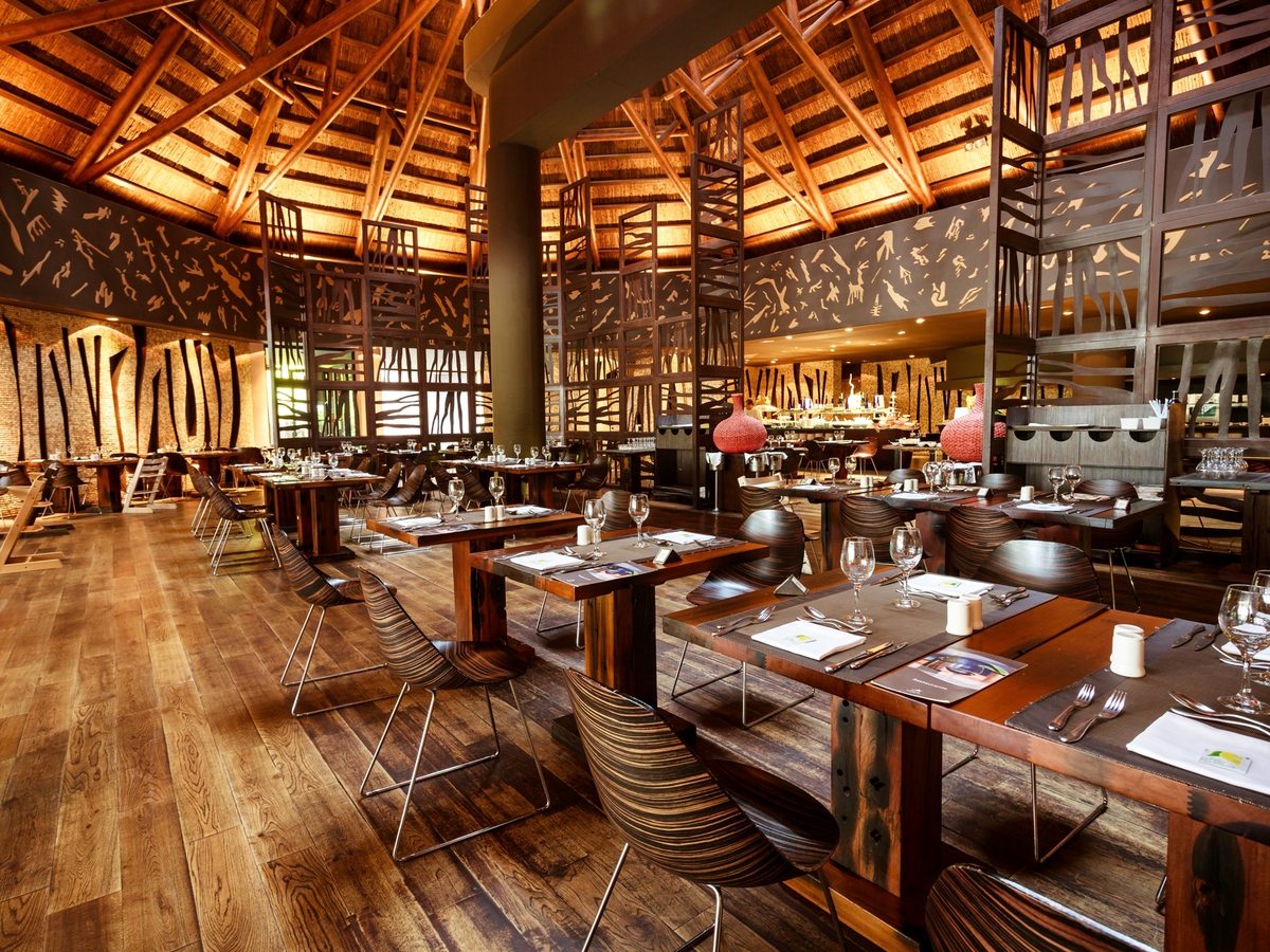 A large restaurant dining room under a high thatched roof on timber beams, with dark wood tables set for a meal, curved wooden chairs and carved screen panels along the walls.