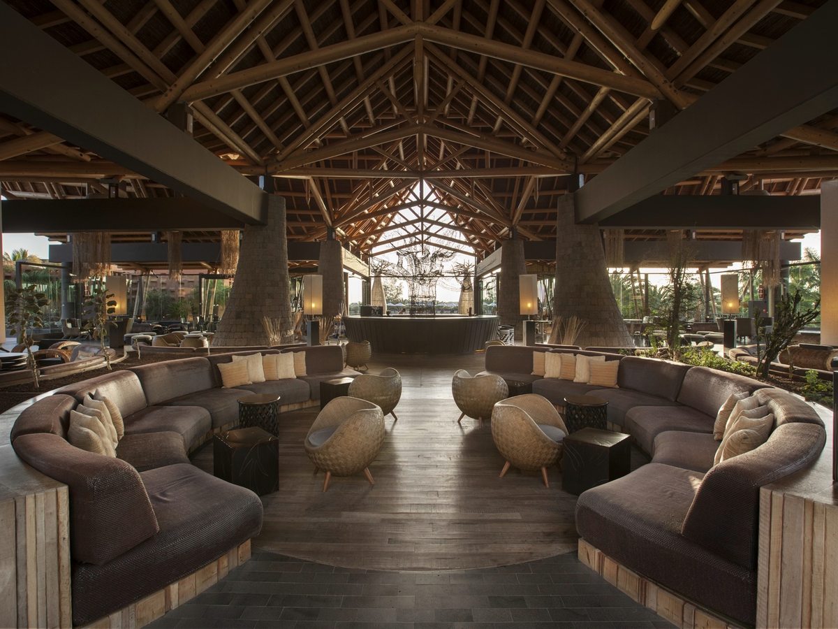 An open-sided lobby lounge with a high thatched roof on timber rafters, ringed by curved sofas and woven armchairs around low tables on a polished floor.