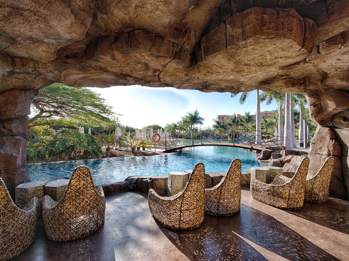 High-backed woven wicker chairs on a stone floor under a wide overhanging rock arch, looking onto a turquoise pool with a timber footbridge and palms.