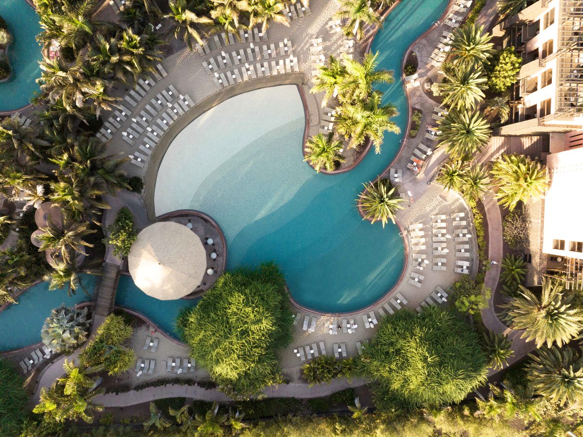 A large freeform pool with a pale shallow shelf at one end deepening to turquoise, a round thatched pool bar at its edge and rows of loungers and parasols on the sand-coloured deck.