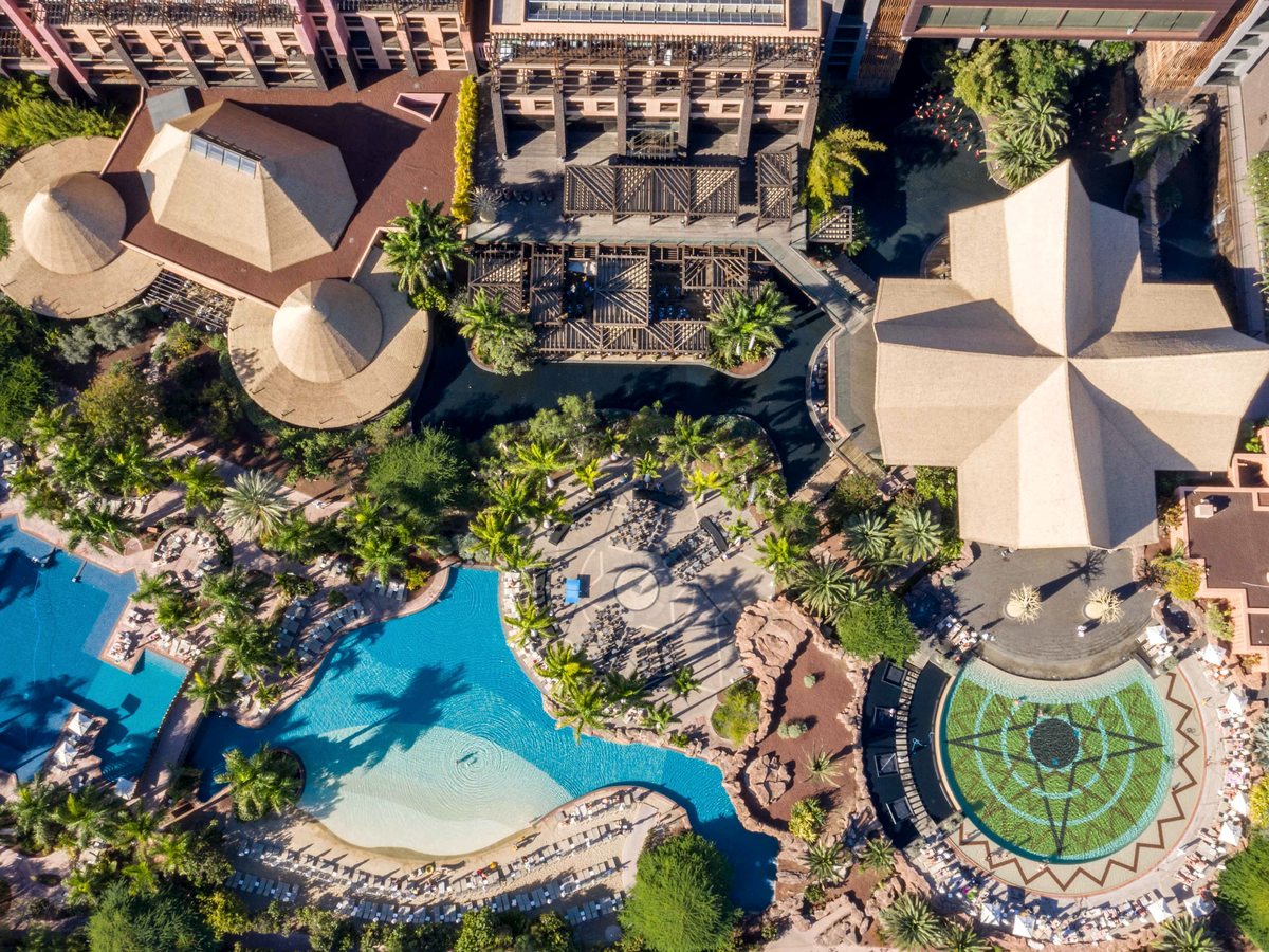 The resort laid out below: thatched conical and star-shaped pavilion roofs, dense palm gardens, a curving blue pool with a pale shallow shelf and rows of sun loungers.