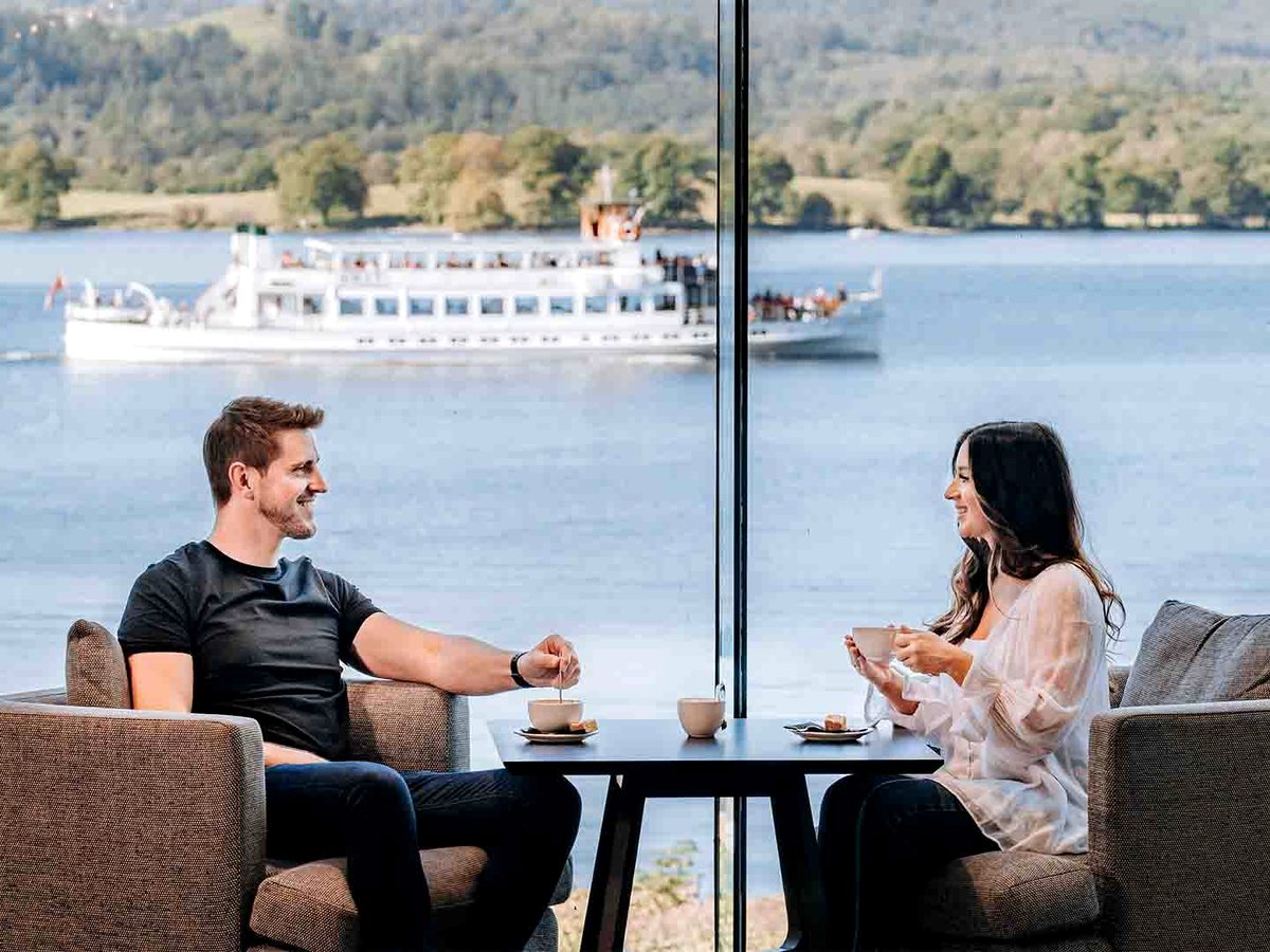 Two guests in armchairs either side of a small black table with coffee cups, at a floor-to-ceiling window as a white cruise boat passes on the lake.