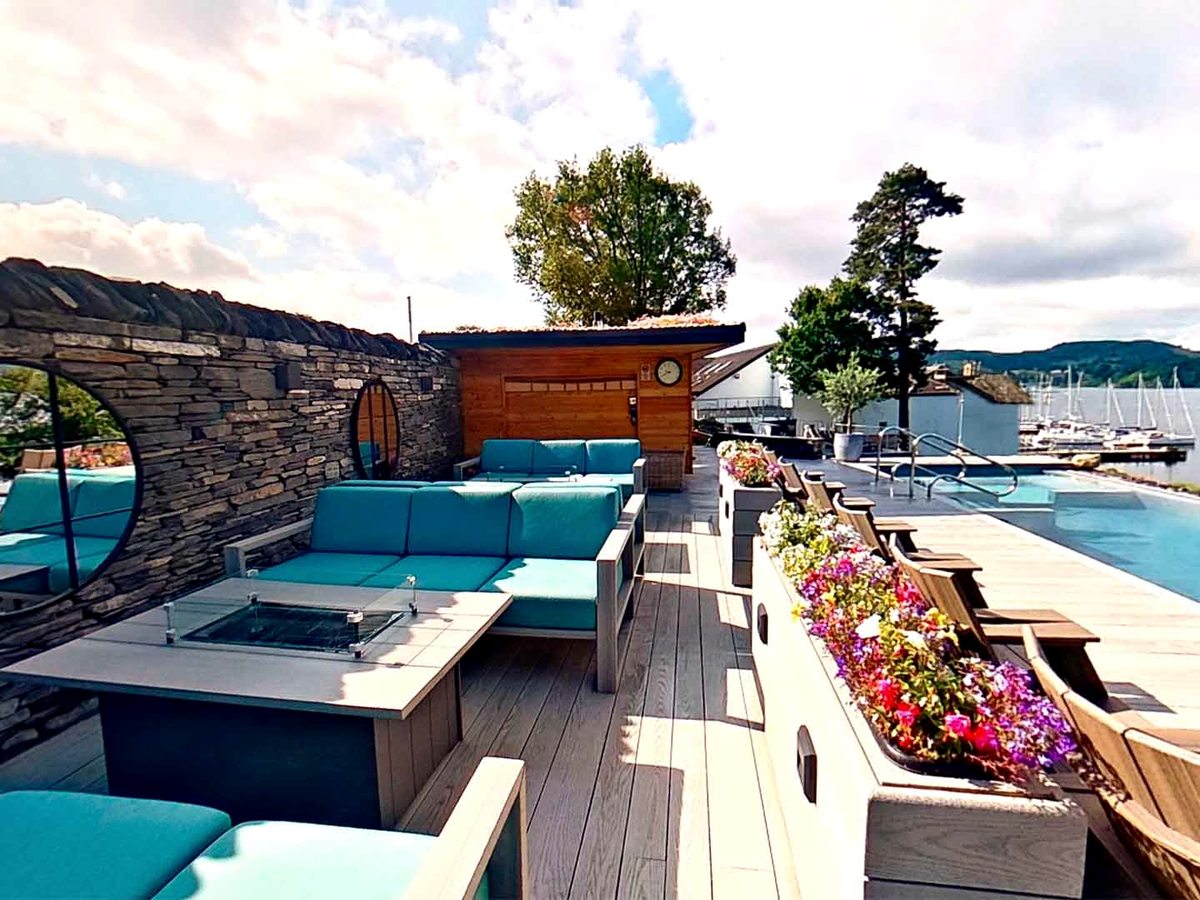 A decked terrace with turquoise sofas around a gas fire pit, a timber sauna cabin set into a slate wall, planters of summer flowers and a pool at the far end.