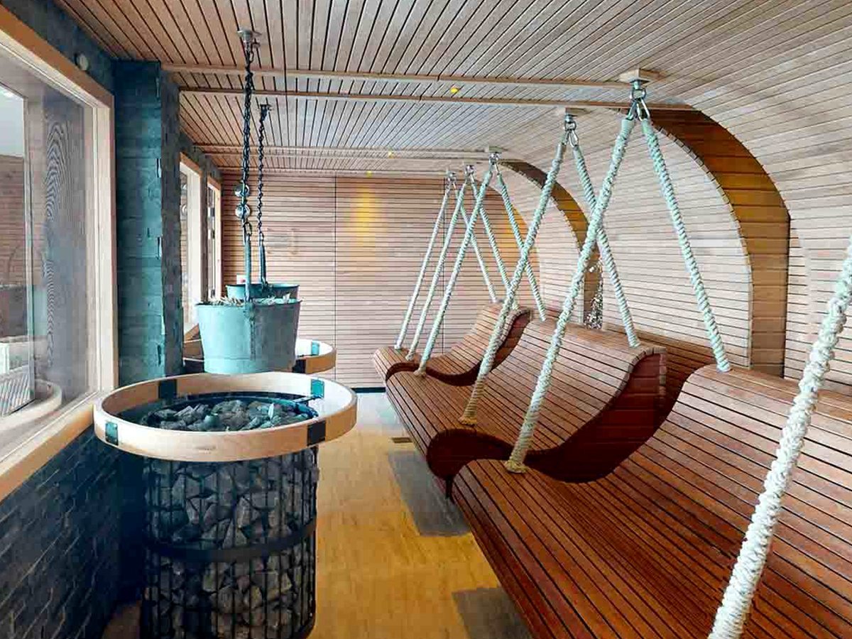 A timber-lined room with contoured wooden swing seats hanging on ropes, and a stone-filled heater with a metal water bucket above it.
