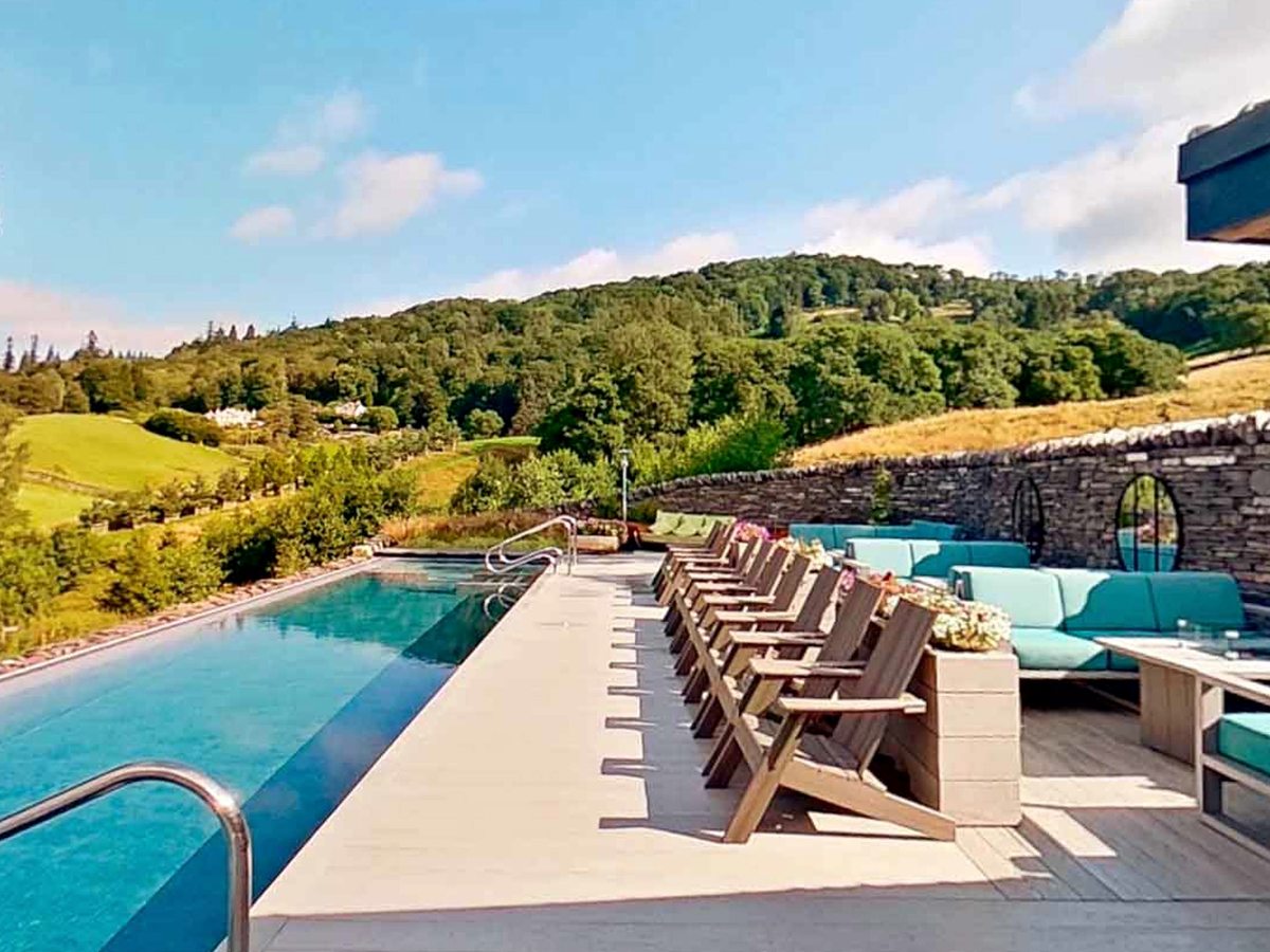 An outdoor infinity pool edged by a row of wooden loungers and turquoise sofas, with wooded fells and fields rising beyond.