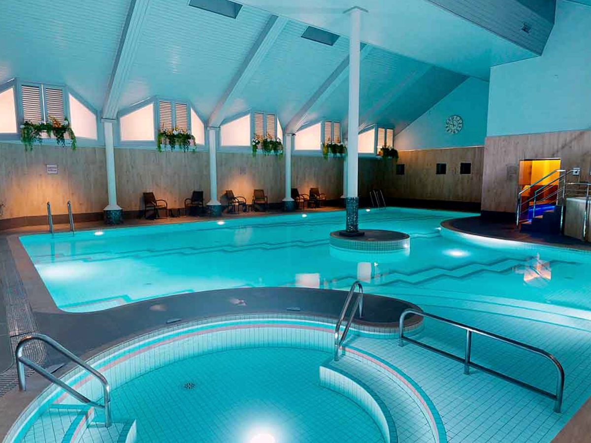 An indoor pool under a white vaulted ceiling with dormer windows and hanging plants, a raised round pool in the foreground and chairs along the far wall.