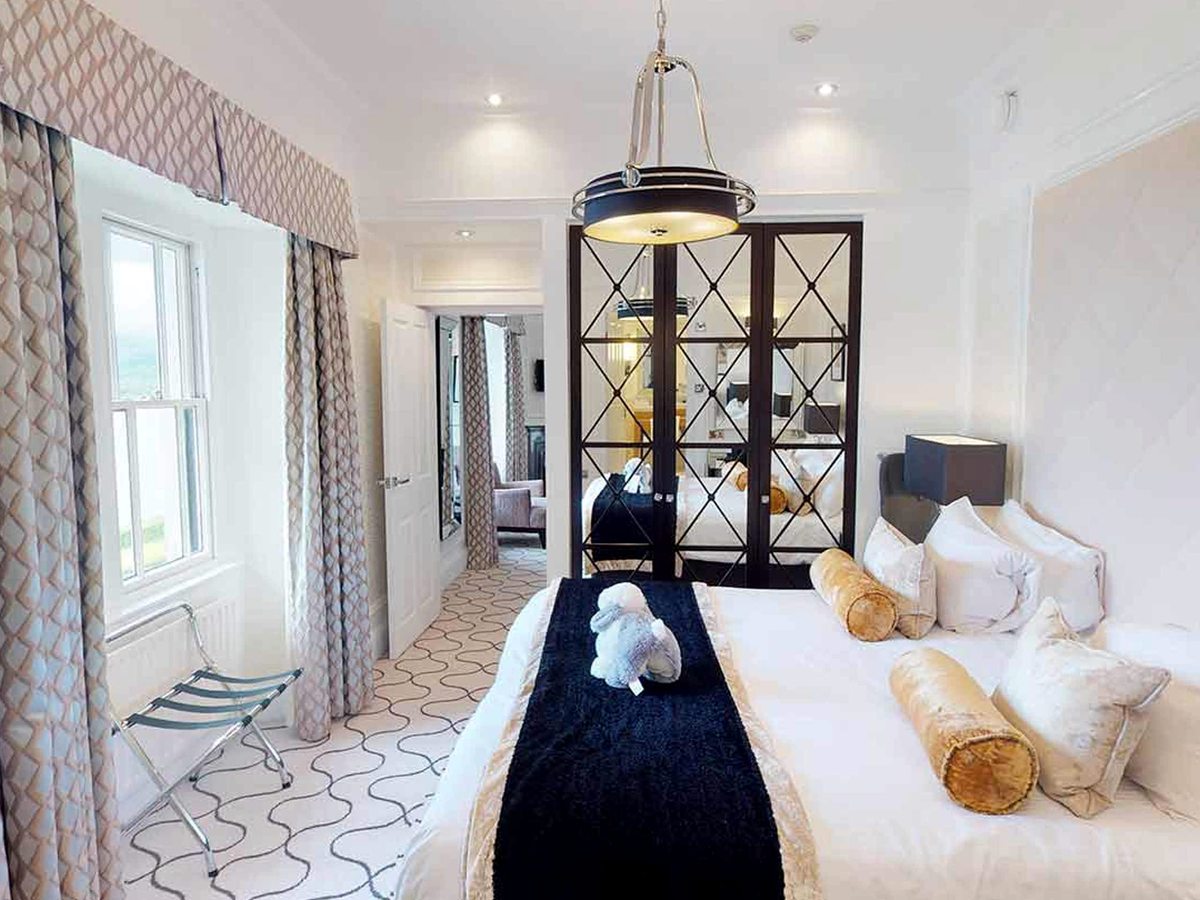 A bed with a navy runner and gold bolsters beside mirrored wardrobe doors, with a sash window on one side and an open doorway through to a sitting room.