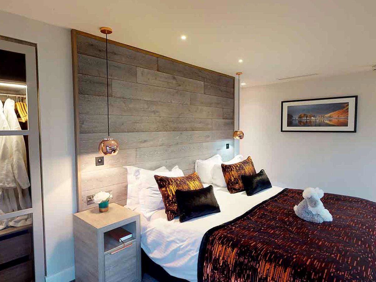 A bed against a grey wood-plank feature wall with copper pendant lights, a bedside cabinet, and an open wardrobe holding white robes.